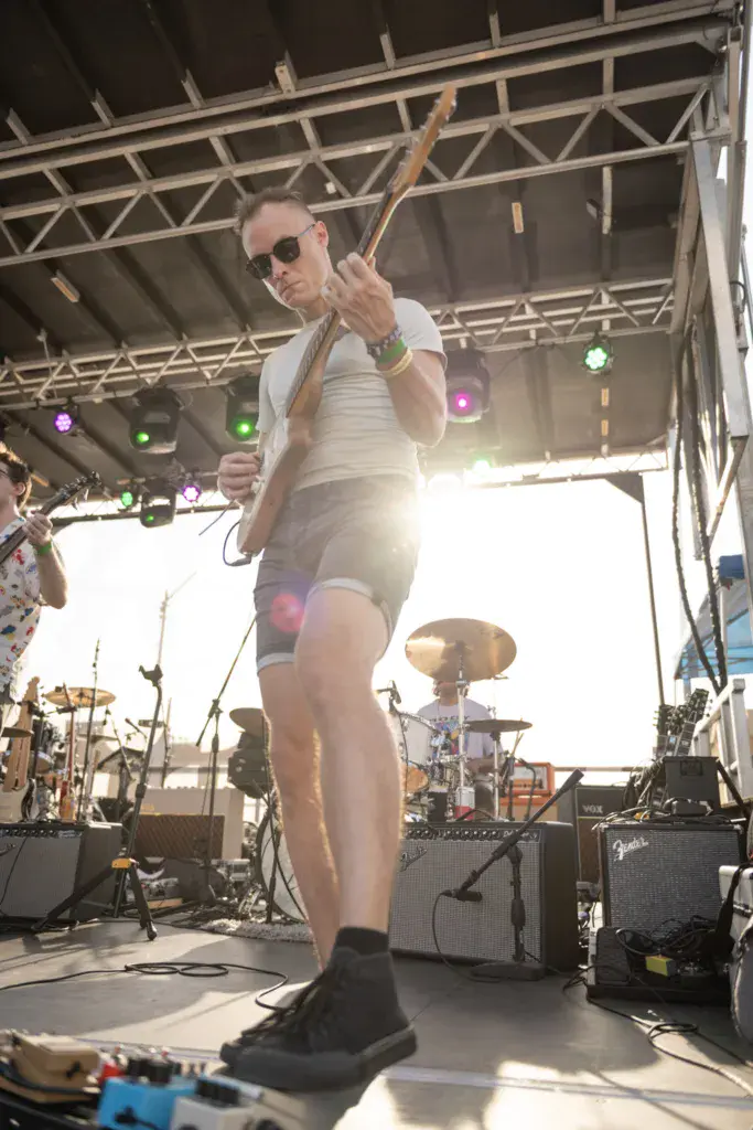 A performer in sunglasses and white tank top plays bass guitar on stage under a metal truss structure during daytime.
