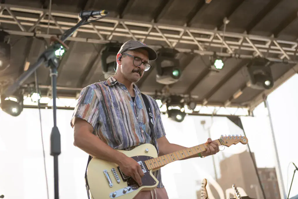 A musician wearing glasses and a striped shirt plays an electric guitar on an indoor stage beneath a metal truss ceiling.