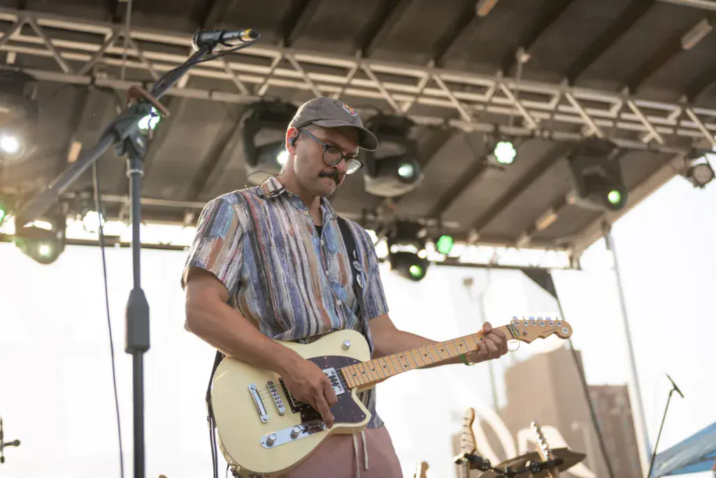 A guitarist wearing a striped shirt and cap performs on stage under a metal roof structure with stage lighting.