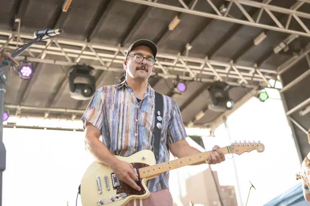 A musician wearing glasses and a striped shirt plays electric guitar on stage under industrial lighting.