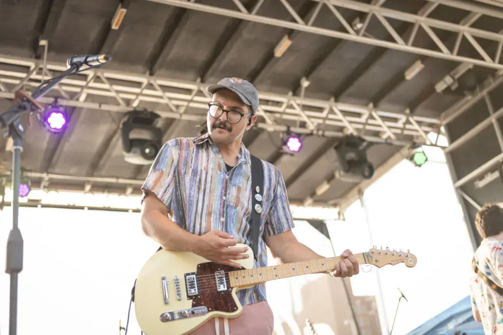 A performer wearing glasses and a striped shirt plays an electric guitar on stage under industrial lighting.