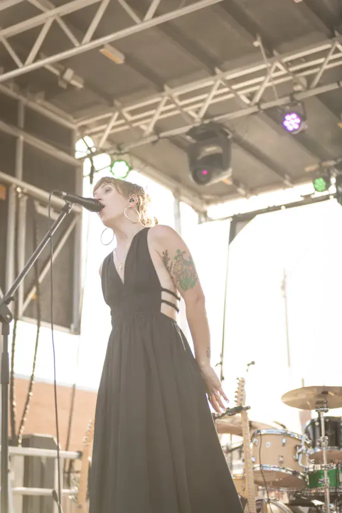 A performer sings into a microphone on an outdoor festival stage with professional lighting overhead.