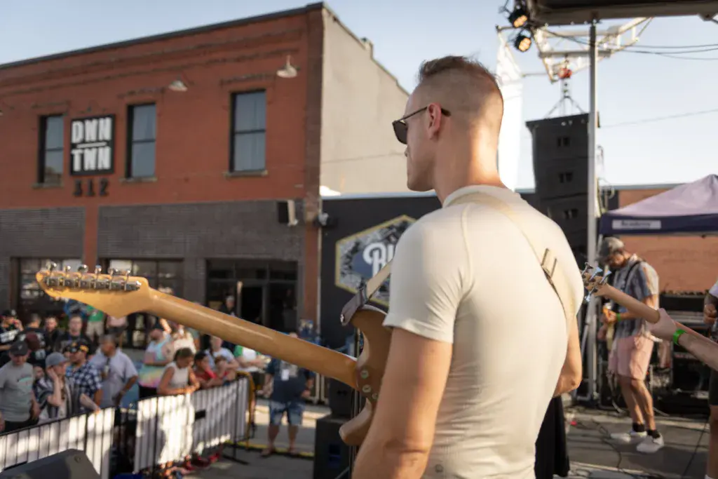 A guitarist wearing sunglasses performs on stage in front of a crowd at an outdoor festival, with brick buildings visible in the background.
