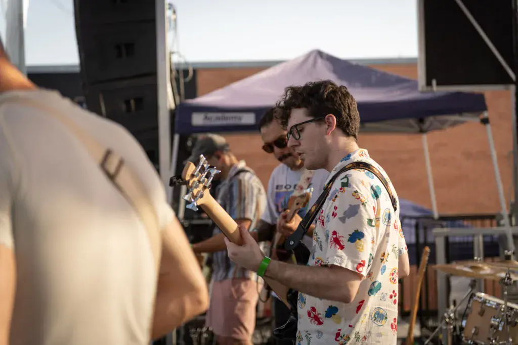 Musicians perform on an outdoor stage with a purple tent canopy visible in the background.