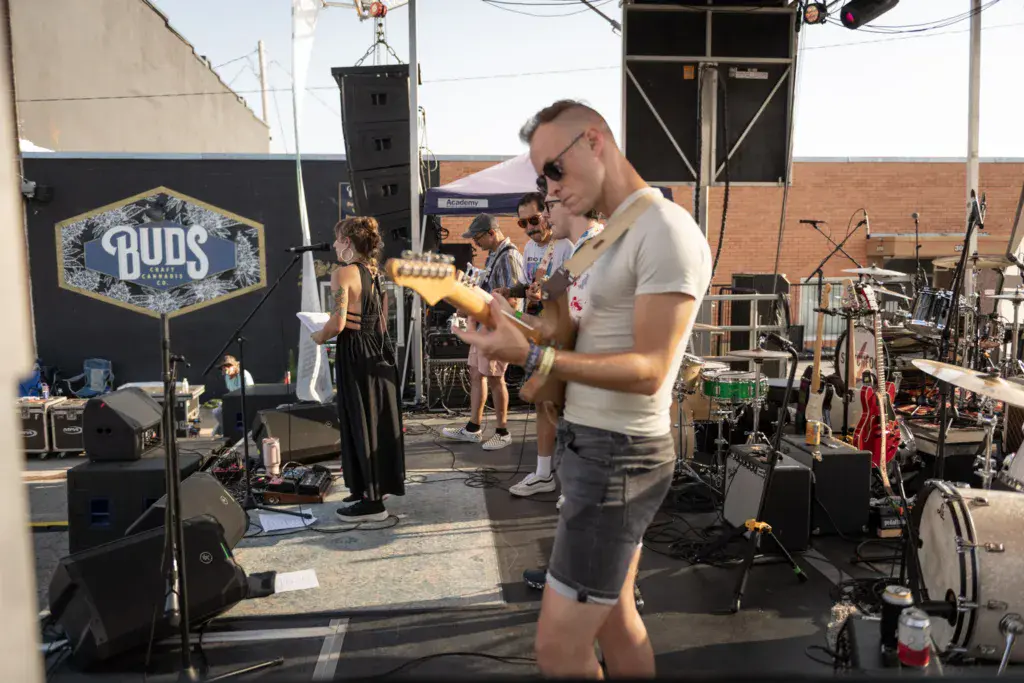 Musician wearing sunglasses plays yellow guitar on outdoor stage with Buds sponsor signage visible