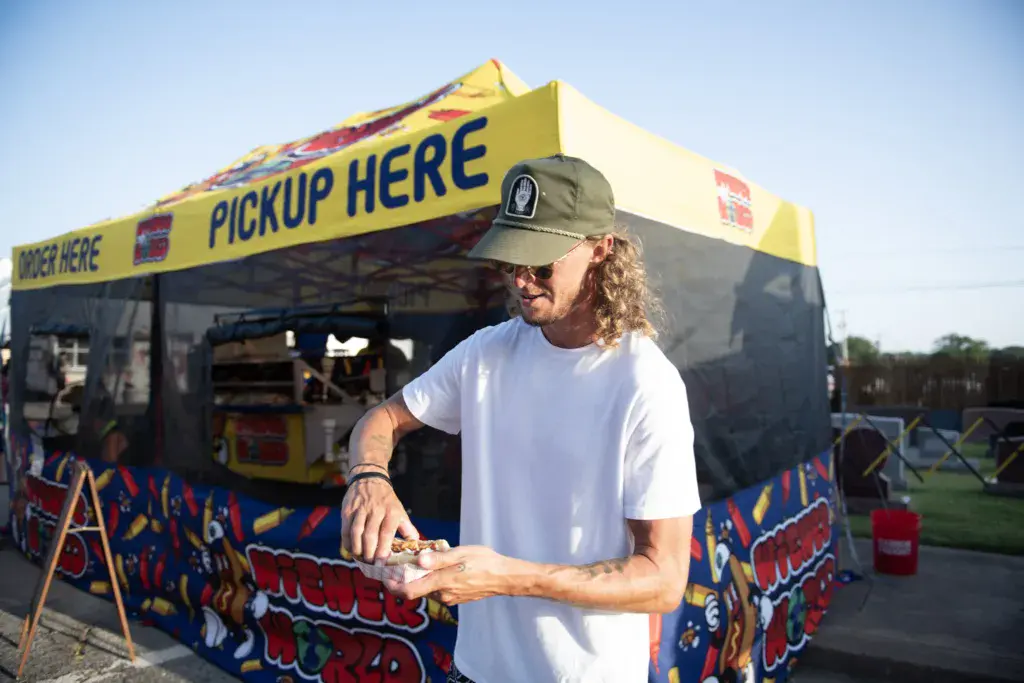 A person holding food stands in front of a yellow and black food vendor booth at an outdoor festival.