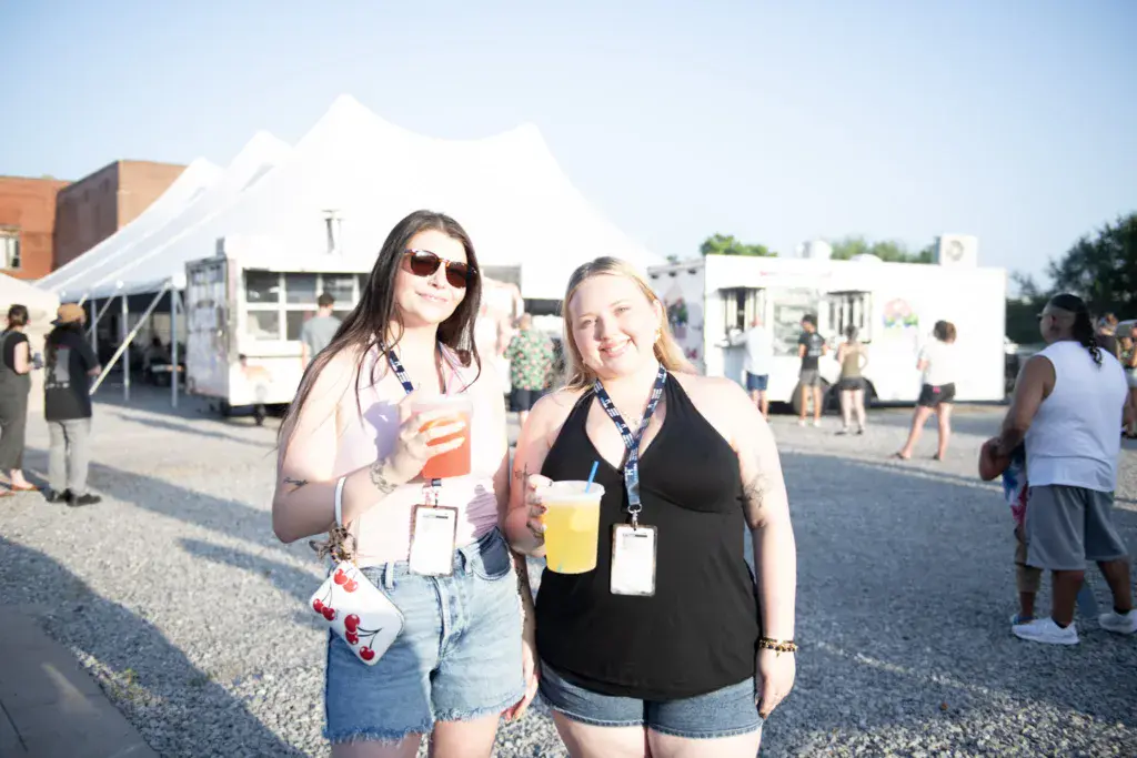 Two festival attendees wearing lanyards and holding drinks smile at the camera in front of vendor tents.
