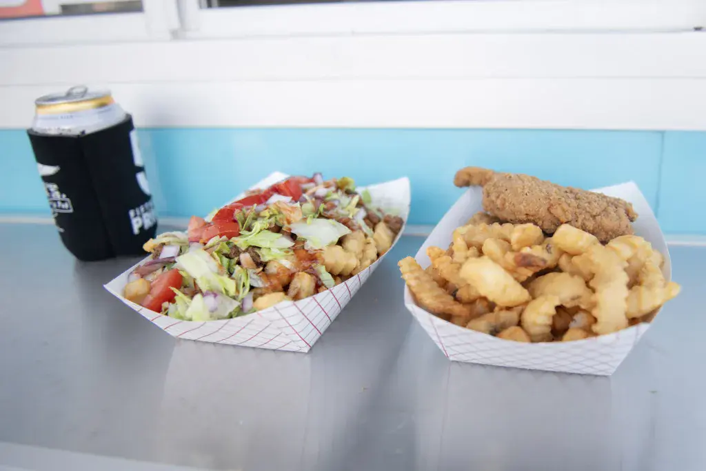 Festival food vendor display with fried chicken, fries, and a salad in paper containers next to a branded beverage holder.