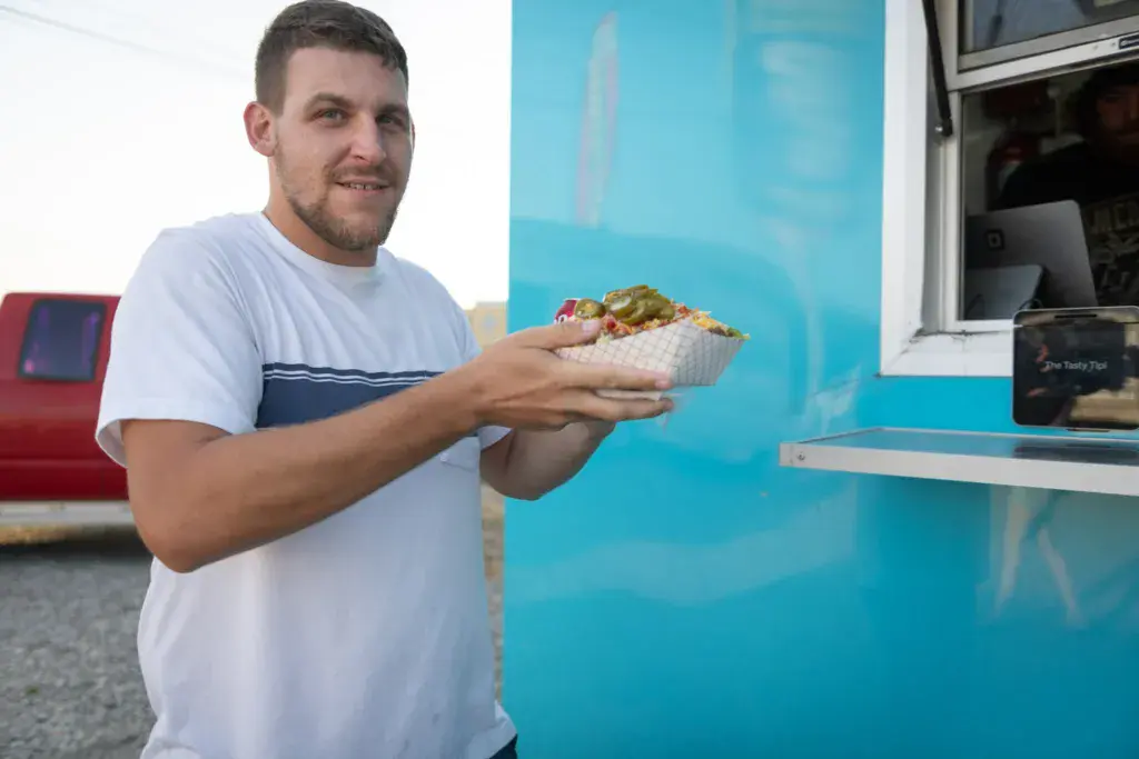 A smiling person holding a food item in a container stands in front of a bright blue food vendor booth at the festival.