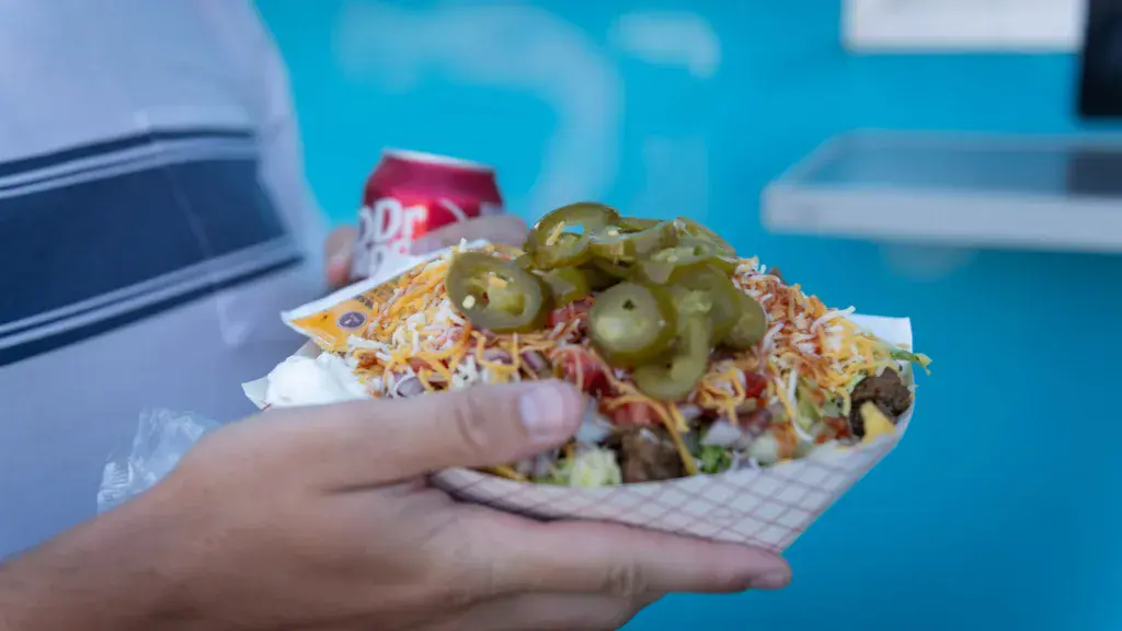A person holds a colorful loaded hot dog with jalapeños and toppings against a bright blue festival background.