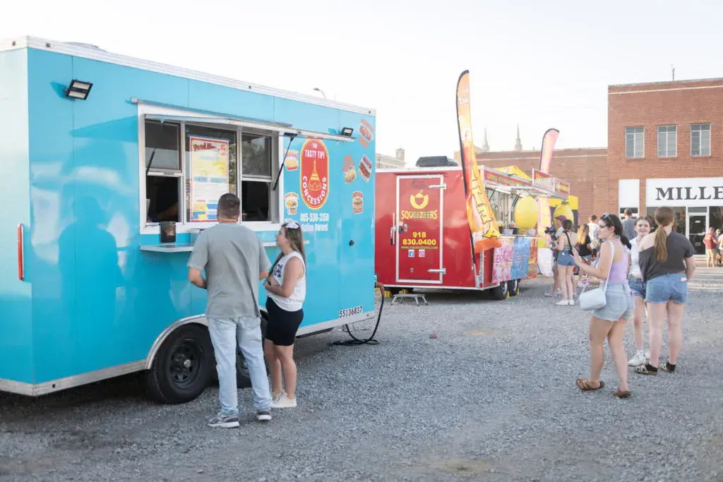 Festival attendees browse food trucks and vendor stands in an outdoor market area with brick buildings nearby.
