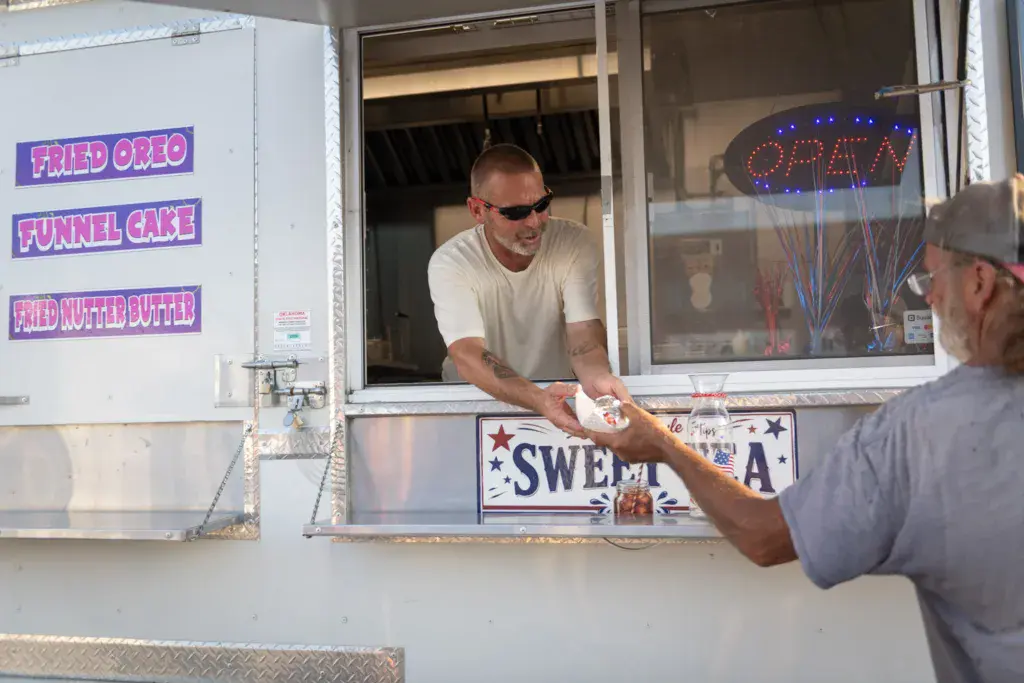 A food vendor hands a paper container to a customer at a festival concession stand.