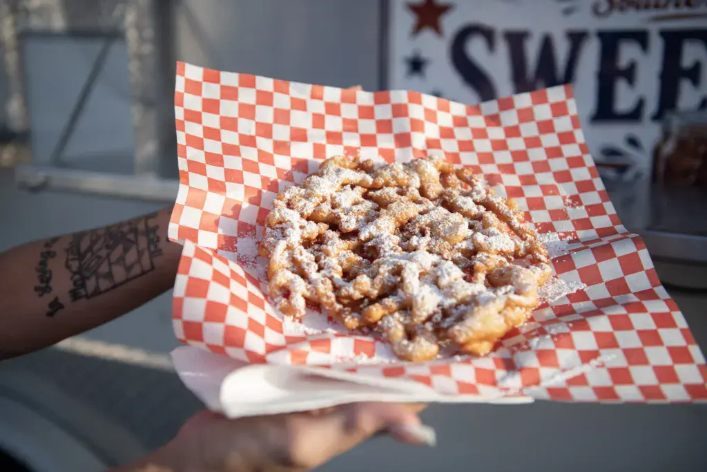 A hand holding a powdered pastry on red and white checkered paper at an outdoor festival.