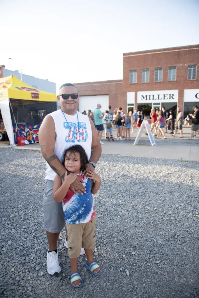 An adult and child pose together in the festival grounds with crowds and brick buildings visible behind them.