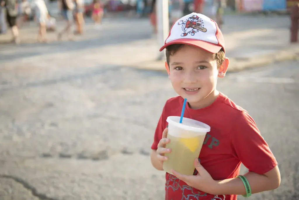 Young child in a red cap and shirt holding a beverage cup at an outdoor festival venue.