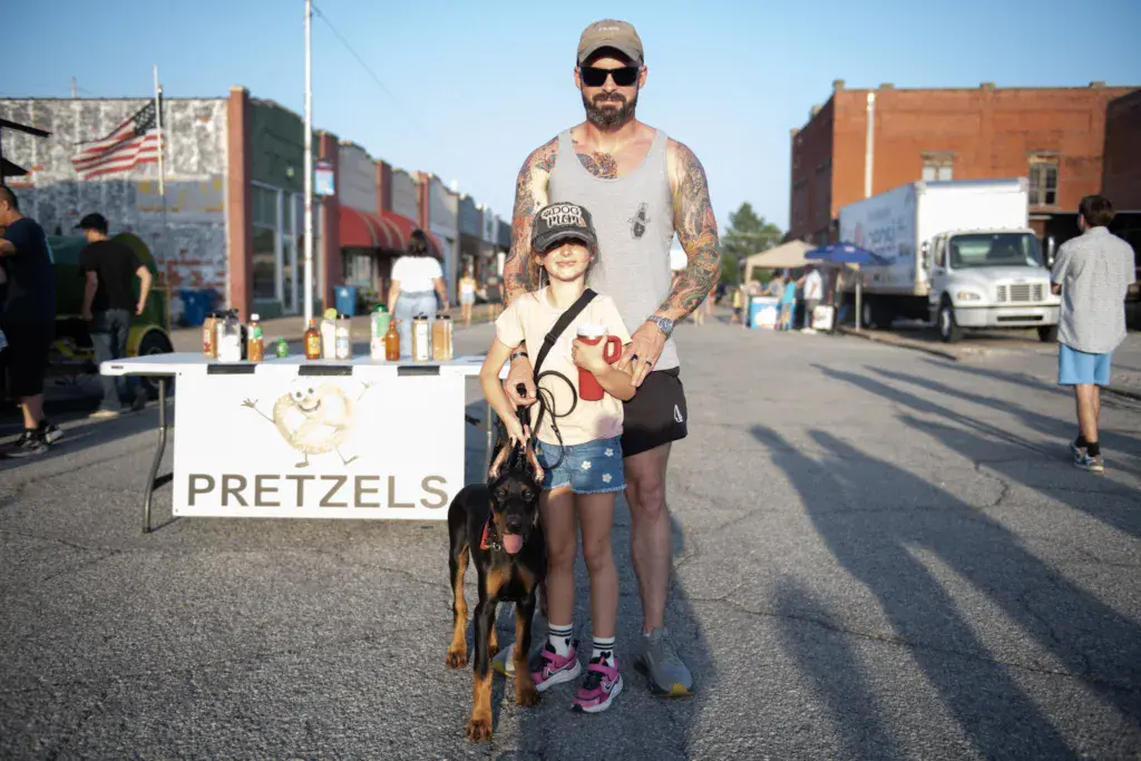 A man with a dog and child pose at a pretzels vendor stand in an urban festival setting.