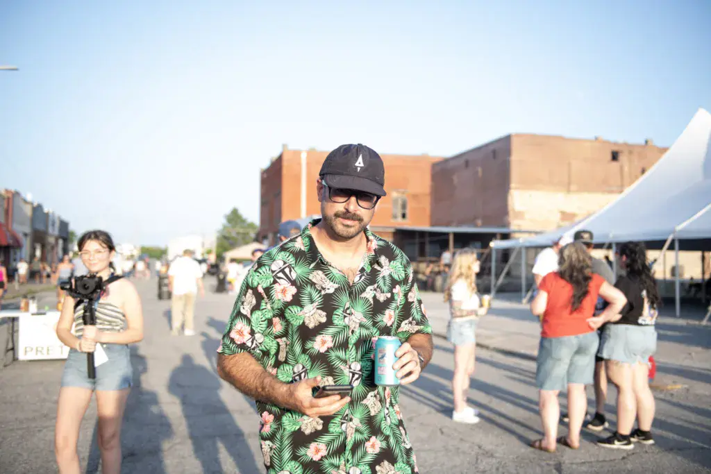 A man in a tropical print shirt and cap smiles at the camera while holding a drink at an outdoor festival with crowds and brick buildings visible in the background.