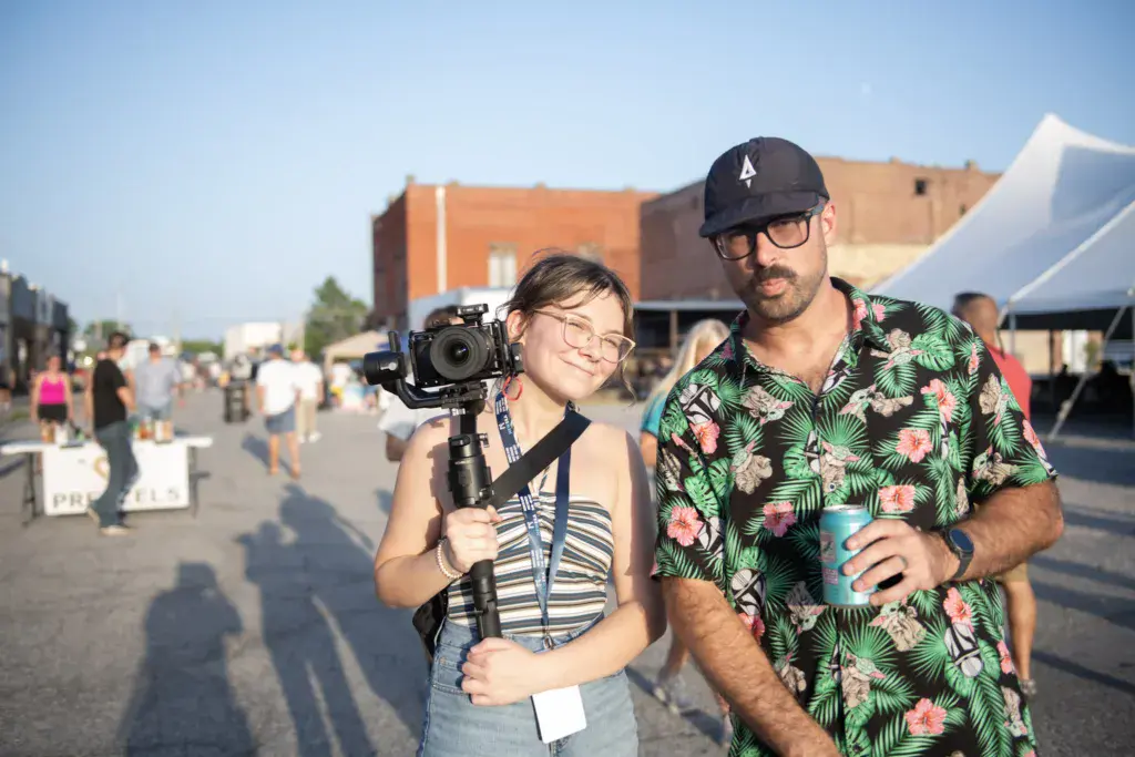 Two people at an outdoor festival, one holding a camera on a gimbal stabilizer while smiling at the camera.