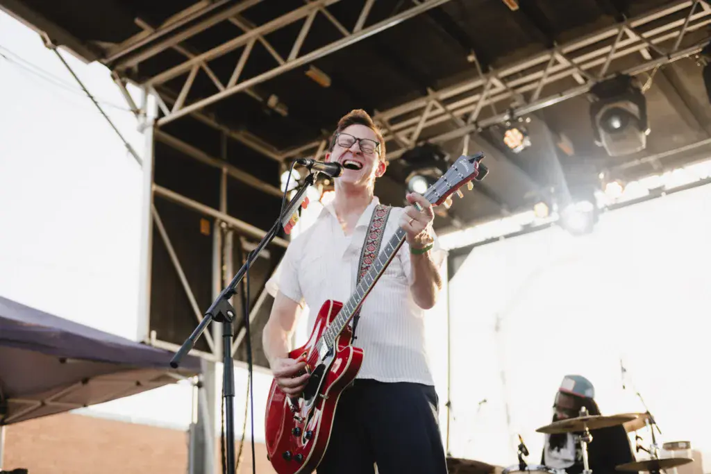 A performer wearing glasses and a white shirt plays a red electric guitar at an outdoor music festival stage.