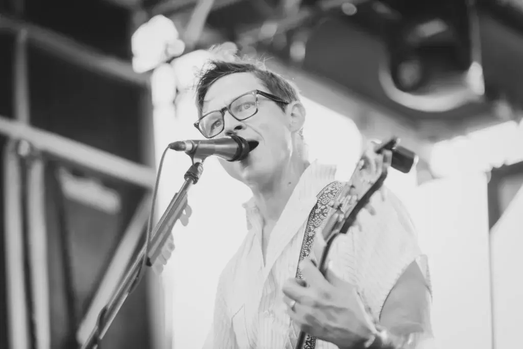 A performer wearing glasses sings into a microphone while playing an electric guitar on stage.