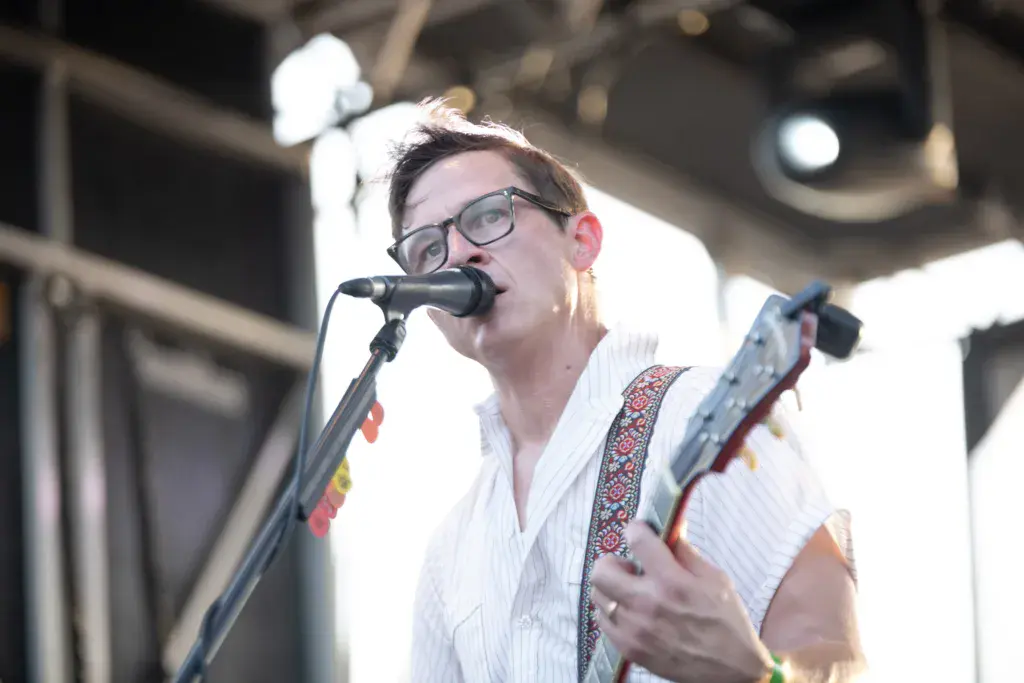A performer wearing glasses and a white shirt sings into a microphone while playing an electric guitar on stage.
