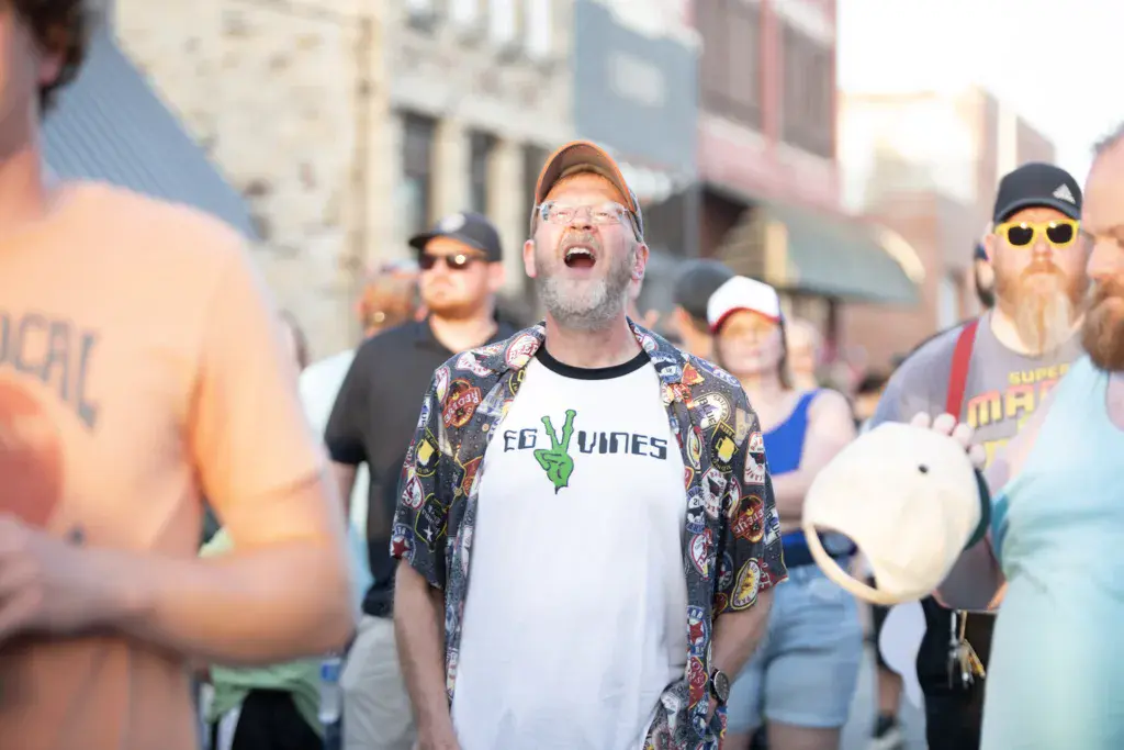 A crowd of festival-goers gathered outdoors, with a man in the center wearing glasses and a colorful shirt laughing with his mouth open.