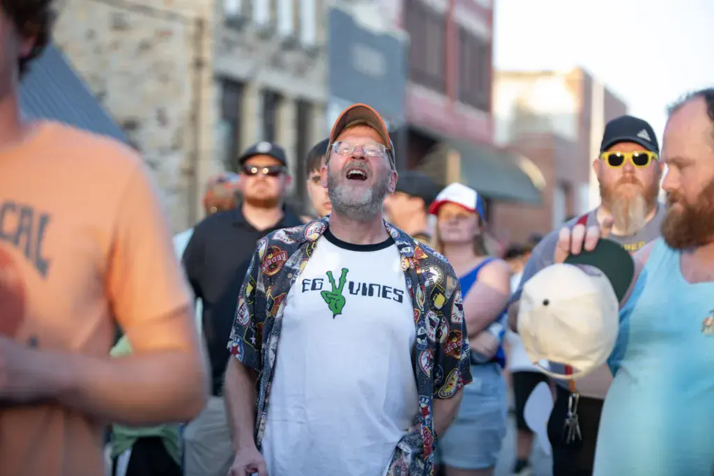 A man in glasses and a patterned shirt sings enthusiastically among a crowd of festival attendees in an urban setting.