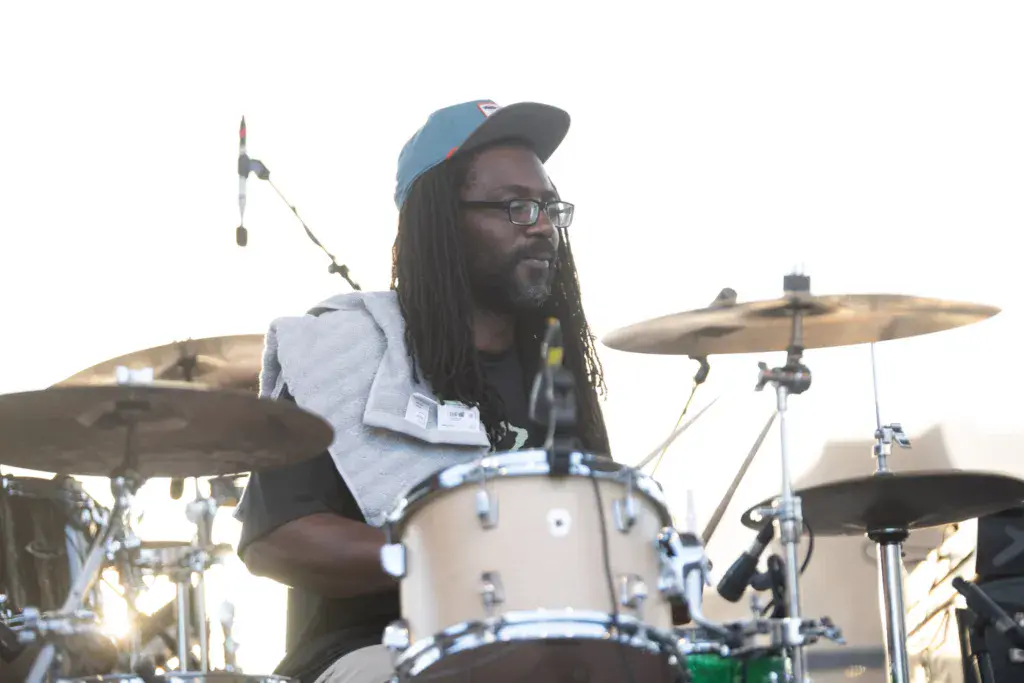 A drummer wearing glasses and a cap performs at an outdoor music festival with cymbals and drum kit visible.