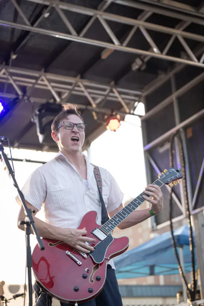A guitarist wearing glasses and a light pink shirt plays a red semi-hollow body electric guitar on stage under professional lighting.