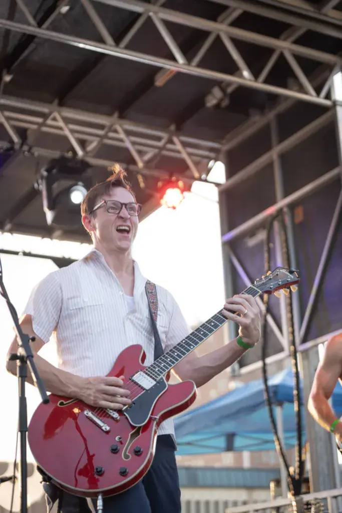 A guitarist wearing glasses and a white shirt plays a red semi-hollow body electric guitar on stage under metal truss lighting.