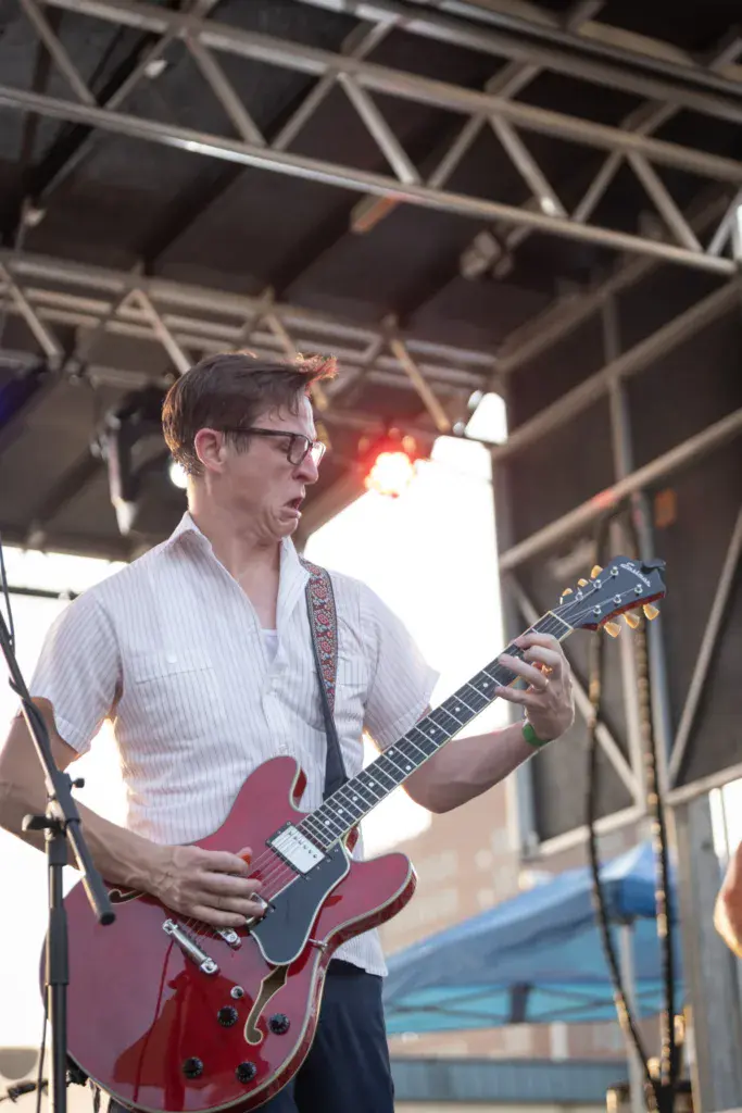 A guitarist wearing glasses and a pink striped shirt performs on stage with a red semi-hollow body electric guitar.