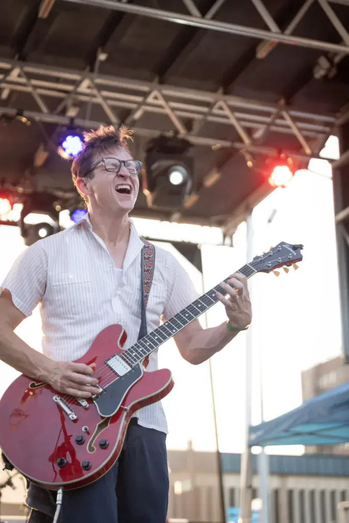 A musician wearing glasses and a light shirt plays a red semi-hollow electric guitar on stage under festival lighting.