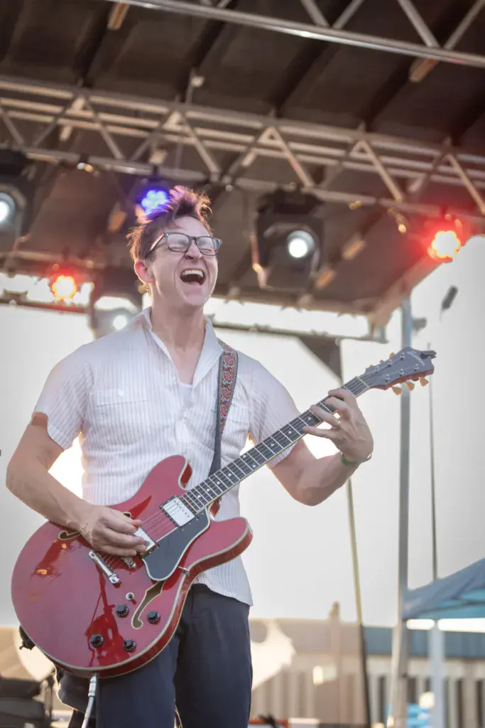 A guitarist performing on stage wearing glasses and a white shirt, playing a red semi-hollow body electric guitar under stage lighting.