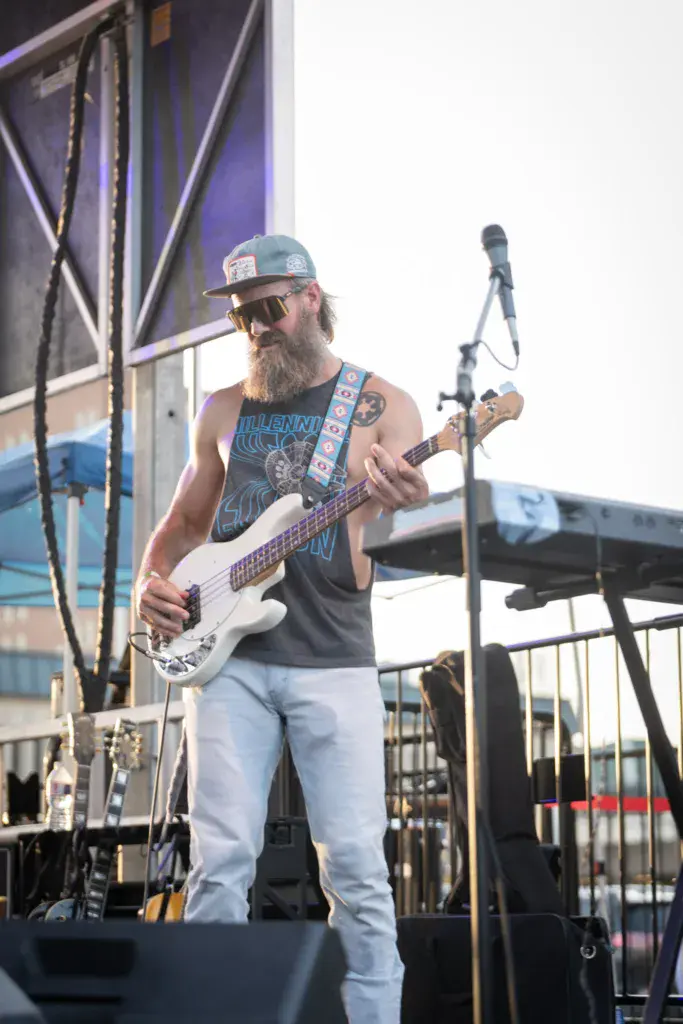 A bass guitarist with a beard and cap performs on stage at an outdoor music festival.