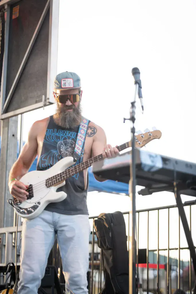 Bearded bass player with long beard wearing sunglasses and cap performs on stage with microphone.