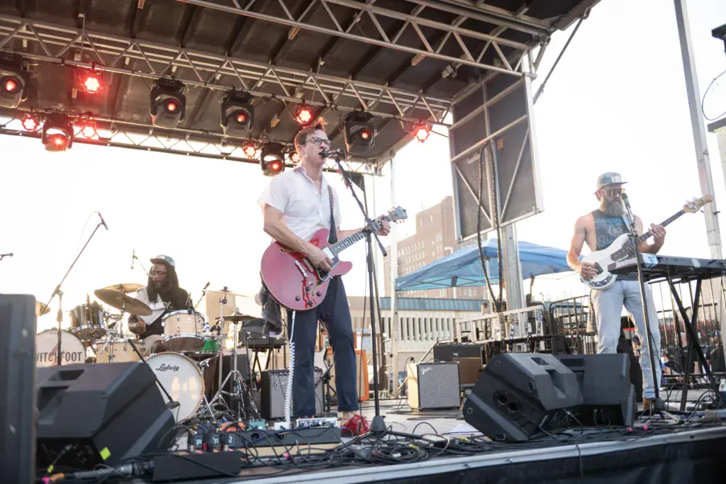 A rock band performs on an outdoor stage with red stage lighting, featuring guitarist, drummer, and bassist.