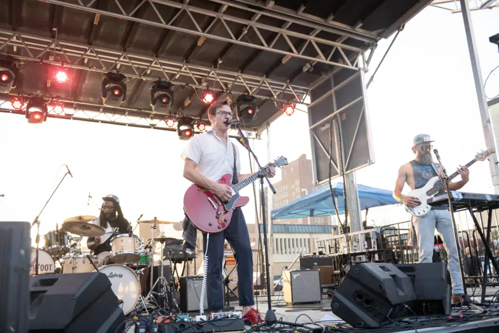 A band performs on an outdoor festival stage with a drummer, guitarist, and bass player under professional lighting.