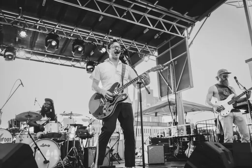 A band performs on an outdoor stage with a guitarist singing lead vocals, drummer, and bass player visible under a metal-frame canopy.