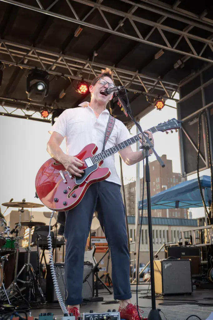 A musician with glasses plays a red semi-hollow body electric guitar and sings into a microphone on an outdoor festival stage.