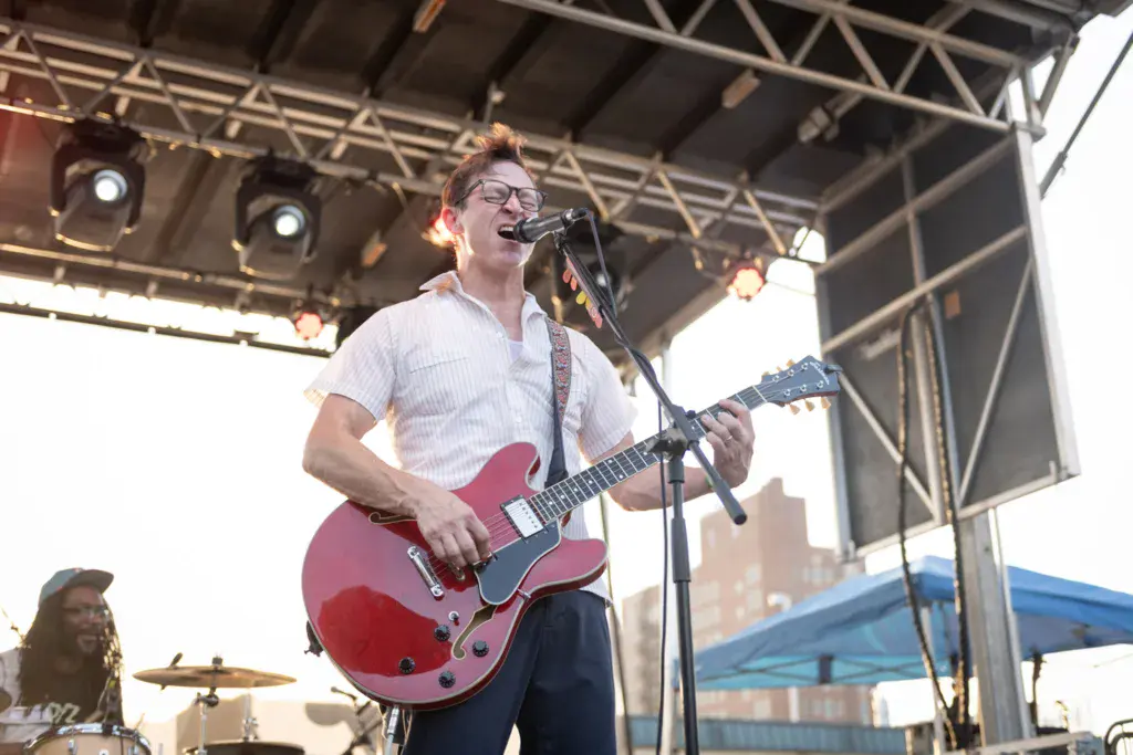Musician performing on stage with a red guitar at an outdoor music festival under a metal roof structure.