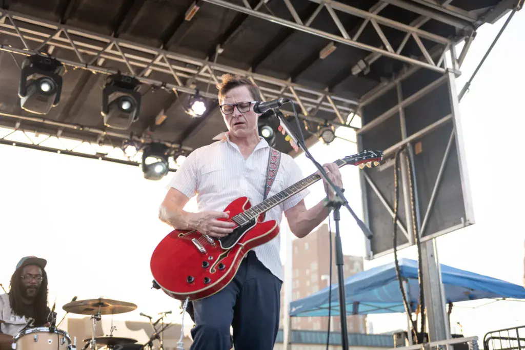 Musician with glasses playing red electric guitar on stage under metal roof structure with stage lights.