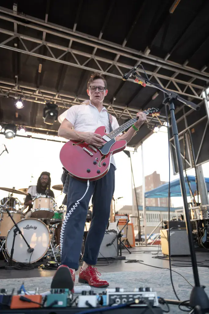 Guitarist in white shirt and glasses performing on outdoor festival stage with drummer visible in background.