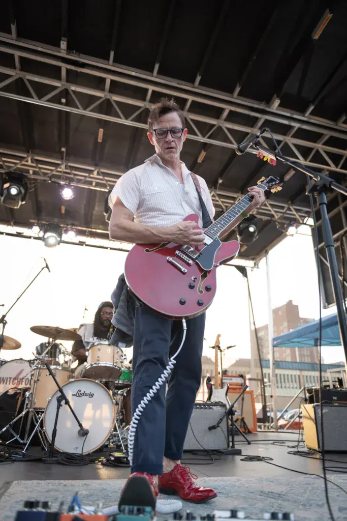 Guitarist in white shirt and red shoes performs with purple semi-hollow body guitar on outdoor festival stage.