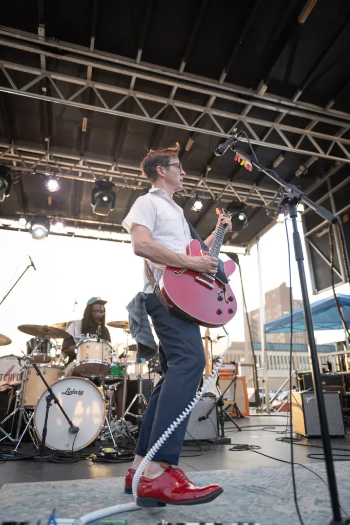 Guitarist in white shirt and red shoes performs with band on outdoor festival stage under industrial truss lighting.