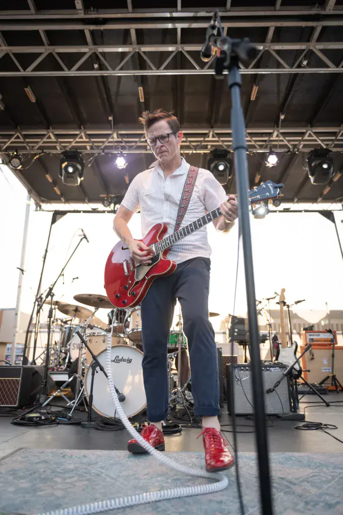 Guitarist wearing glasses and red shoes performs with a red semi-hollow body electric guitar on a festival stage.