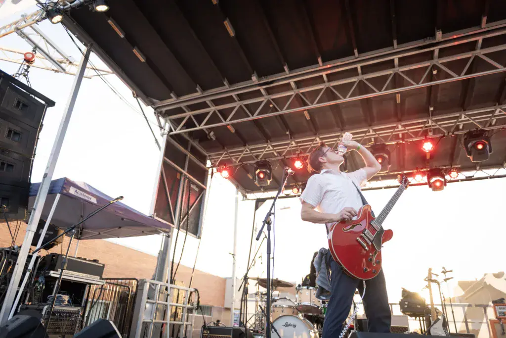 A guitarist performs on stage under a metal truss structure with red stage lights during an outdoor music festival.