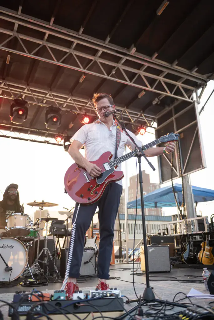 A guitarist performing on stage under a metal canopy with red stage lights and a drummer visible in the background.