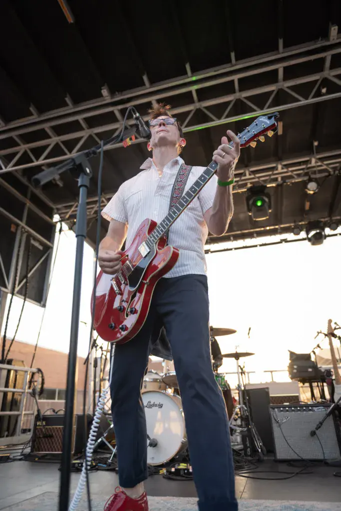 A guitarist wearing glasses and a white shirt plays a red semi-hollow body electric guitar on stage.
