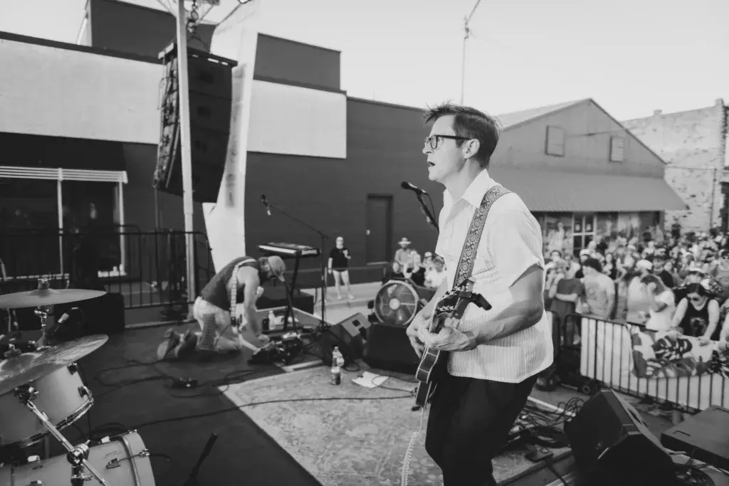 Guitarist performing on stage at an outdoor music festival with crowd watching behind barriers.