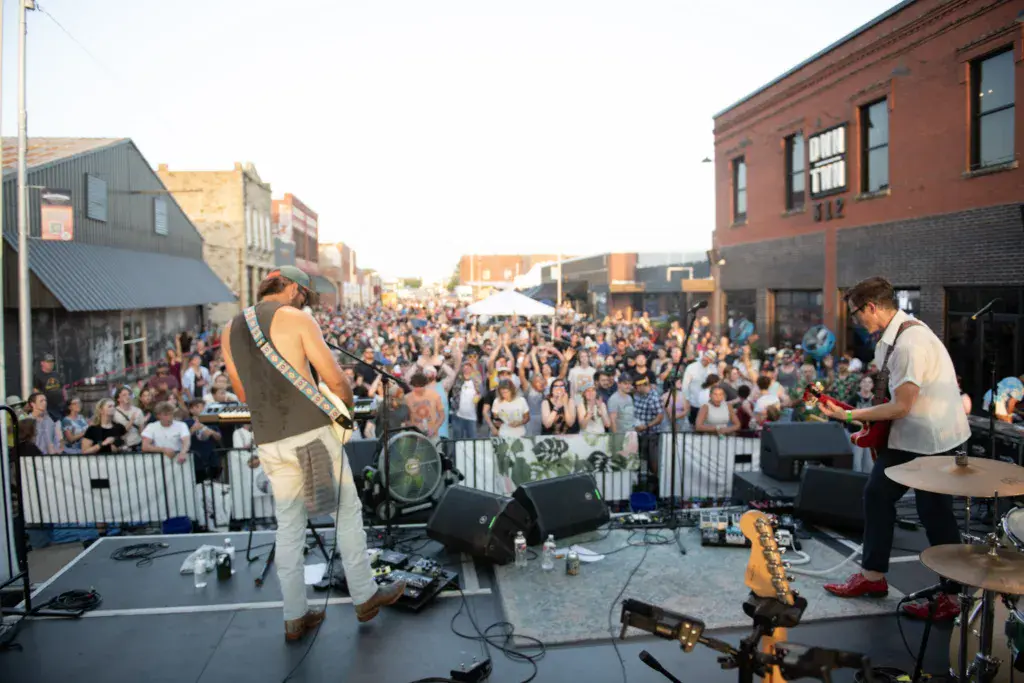 A band performs on an outdoor stage in front of a large crowd in an urban street venue during daytime.